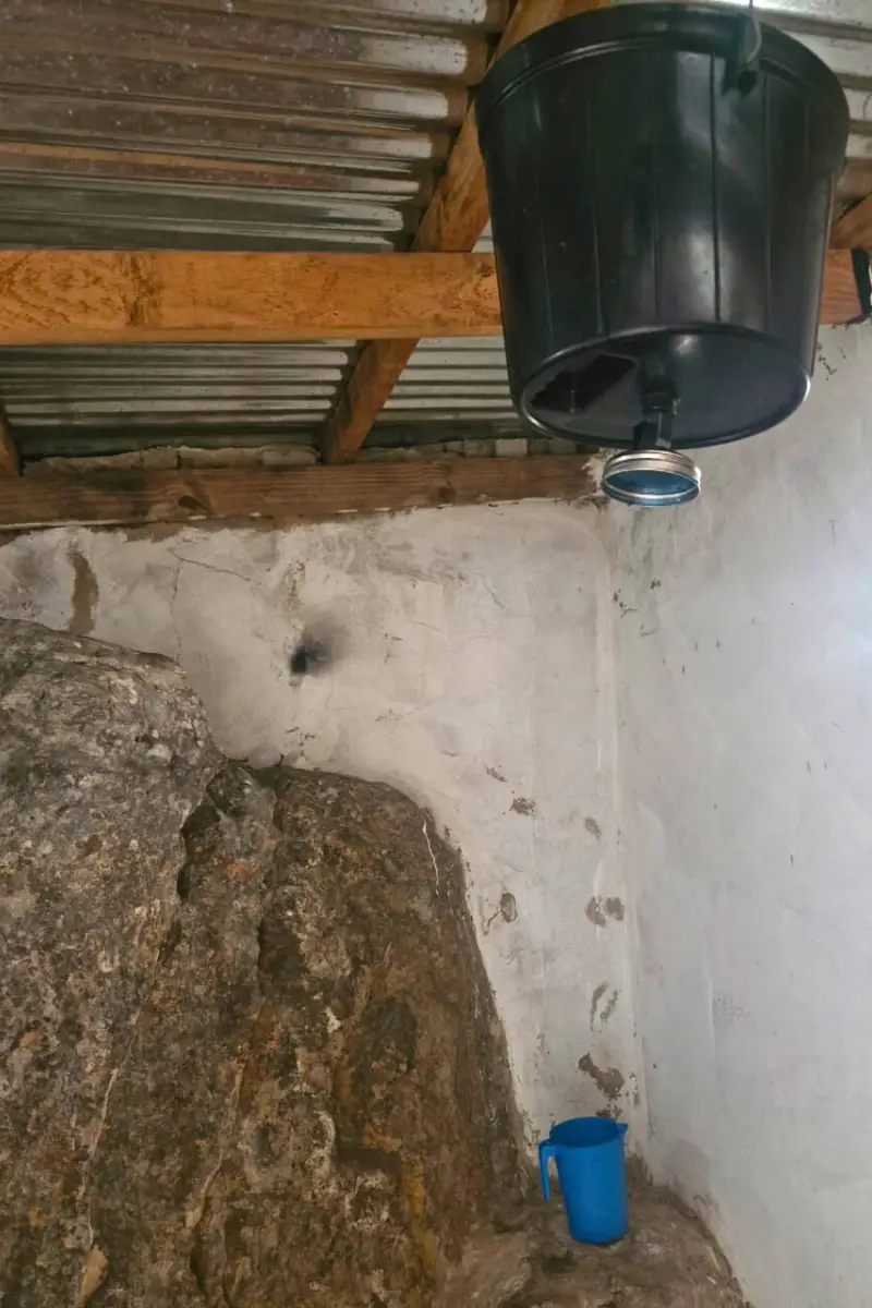Black bucket with shower head suspended from corrugated roof, dripping hot water inside a stone-walled bush shower on the Wild Coast