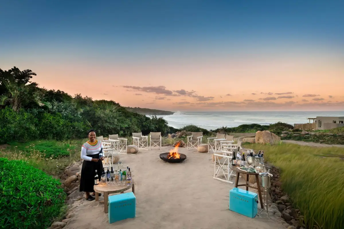 GweGwe Beach Lodge staff setting up outdoor fire pit seating area at dusk overlooking the Wild Coast shoreline