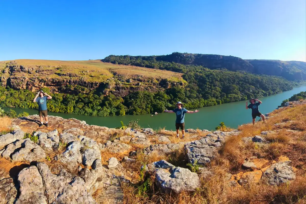 Three hikers with arms outstretched on rocky viewpoint above the lush green Mtentu River gorge, Wild Coast South Africa