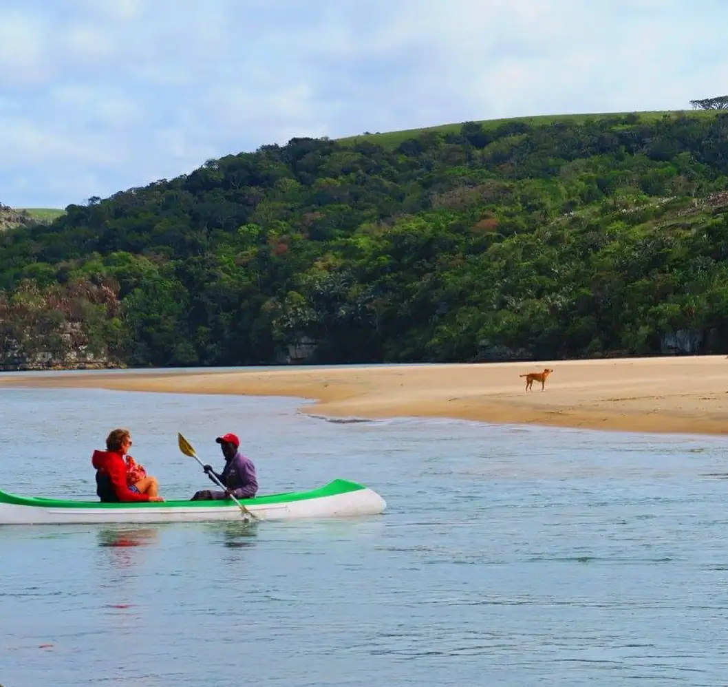 Two hikers paddling kayak across calm Mtentu River with green forested hills and sandy bank behind