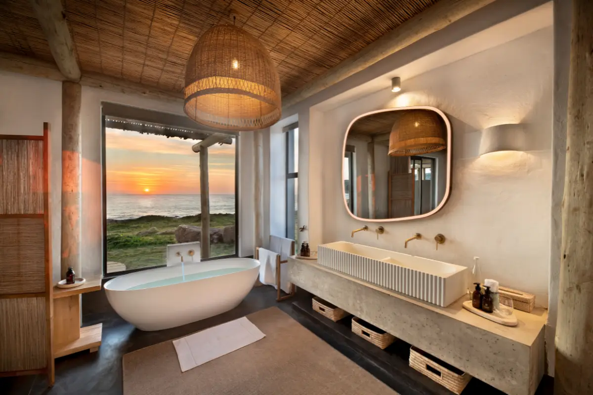Luxury suite bathroom with freestanding oval bath and floor-to-ceiling window framing a Wild Coast sunset at Gwegwe Beach Lodge