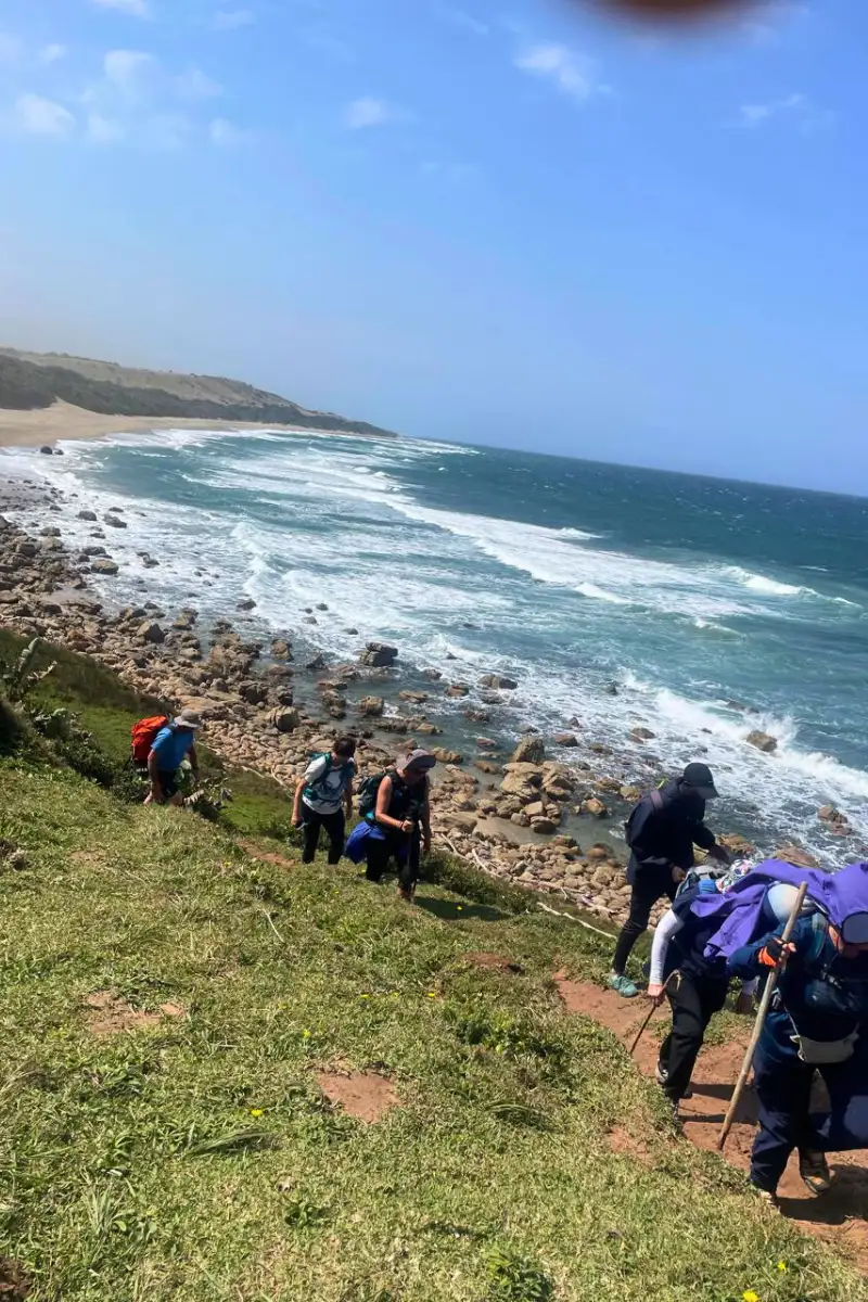 Hikers climbing a grassy coastal headland above rocky shores and crashing Indian Ocean waves on the Wild Coast