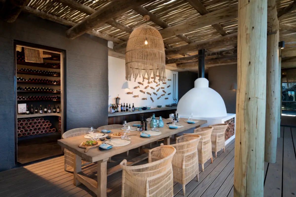 Open-sided dining room at Gwegwe Beach Lodge with long wooden table, woven pendant light, wine cellar and fish-scale wall art