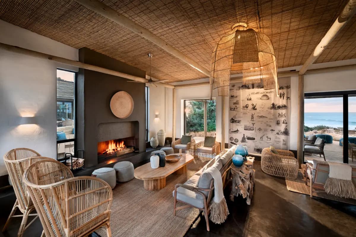 Stylish interior lounge at GweGwe Beach Lodge with wicker chairs, lit fireplace and ocean views through large windows