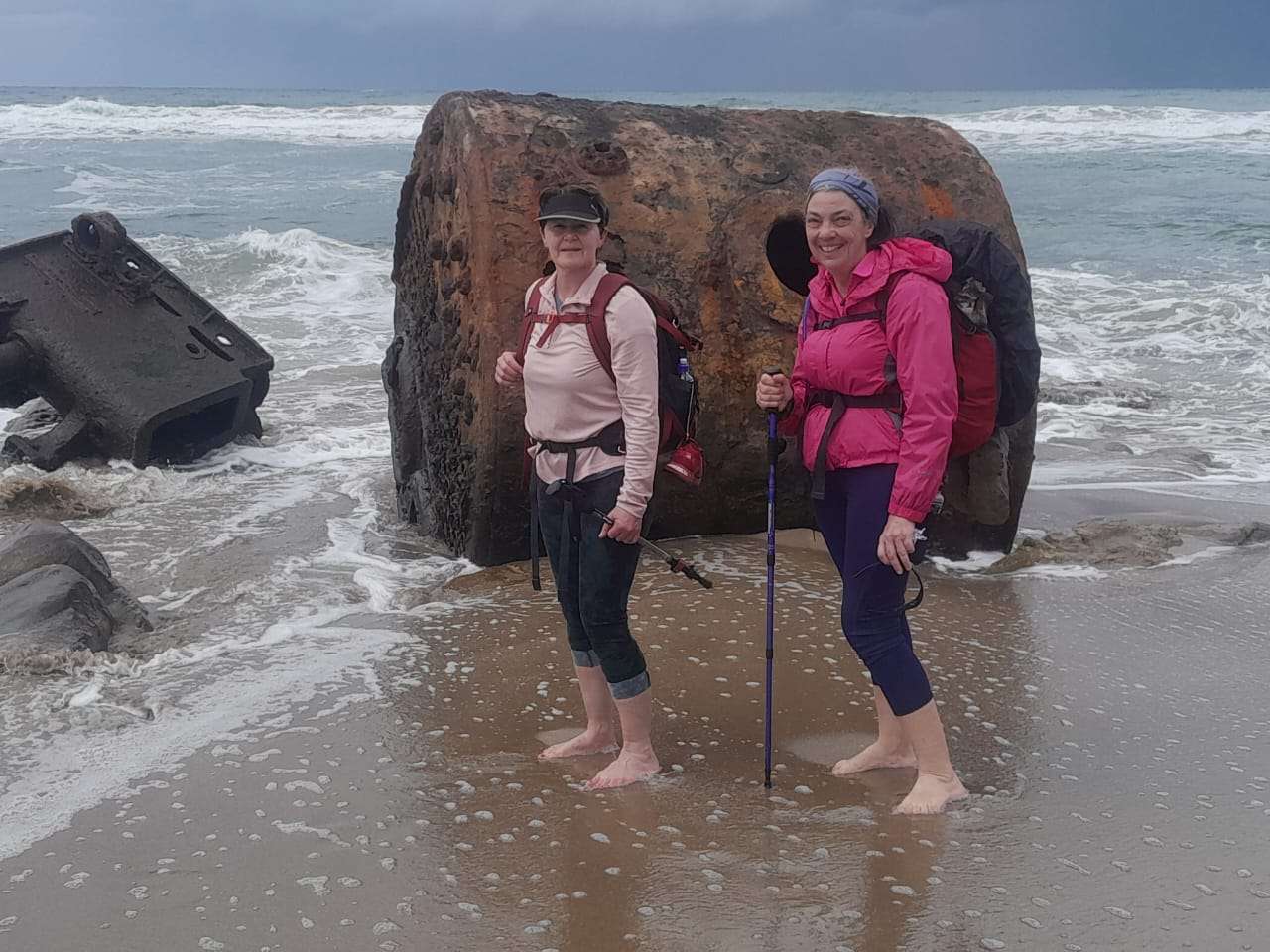 Two hikers standing barefoot beside a massive rusted shipwreck boiler washed ashore on the Wild Coast, South Africa